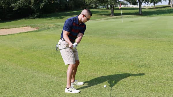 Man in navy shirt, khaki shorts, white shoes, and sunglasses swings a golf club back in preparation of hitting a ball toward a hole in the distance
