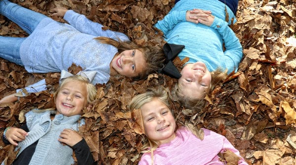 Four young girls lie in fall leaves on ground with their heads pointed toward one another