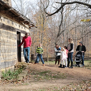 Adults and children stand outside a rustic cabin with leafless trees in the background