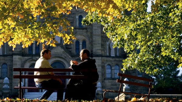 Two men, one in a short-sleeved colored shirt and jeans and one in a monk's robes, sit turned toward one another on a bench under a canopy of colorful fall leaves with a building at Saint Meinrad Archabbey visible in the background