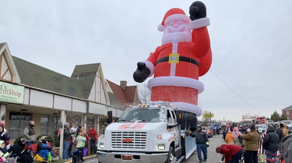 Large inflatable Santa is attached to the back of a truck and drives past storefronts and a crowd of parade-goers