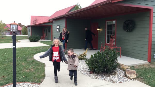 Two children running down sidewalk toward camera with parents in background standing in front of a green cottage with red room at Santa's Lakeside Cottages