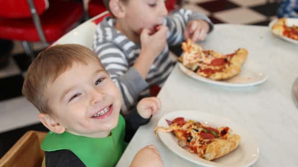 Young boy sits at a table with a slice of pizza on a plate in front of him and smiles at the camera while another young boy sits next to him in the background and eats pizza.
