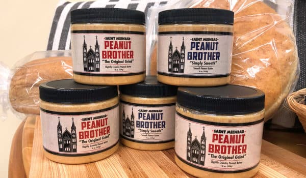 Five jars of Peanut Brother peanut butter are stacked on a wooden cutting board with loaves of bread in clear plastic bags behind them