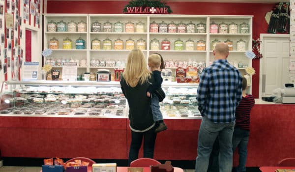A man, woman, and two kids stand in front of a candy counter with shelving featuring jars of colorful popcorn in the background