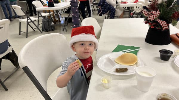 Young boy in gray t-shirt and red Santa hat looks at the camera as he sits at a table in front of a plat of breakfast food