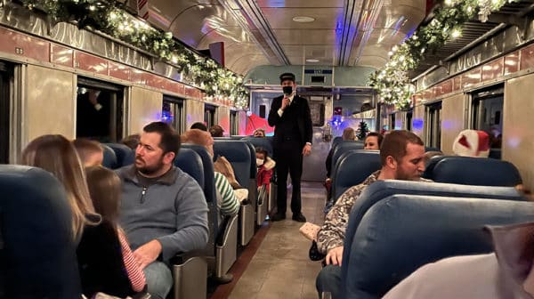 Passengers sit in seats facing forward and backwards inside a train decorated for Christmas as the conductor stands in the aisle 