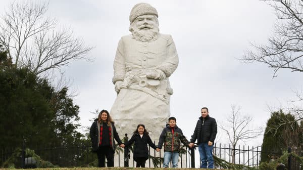 A family of four hold hands and pose in front of a white stone Santa statue at the Santa Claus Museum & Village