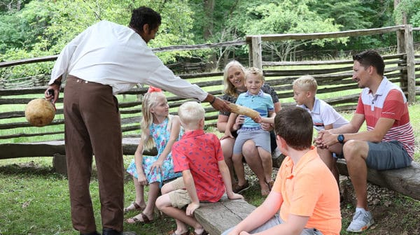 Multiple children and two adults sit on benches in an outdoor space with a split rail fence behind them as a costumed interpreter hands something to one of the boys at the Living Historical Farm at Lincoln Boyhood National Memorial