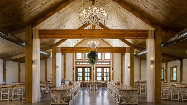 View of inside of large event space, Matilda's Event Barn, shows long tables, wood beams, chandeliers and the front doors and windows