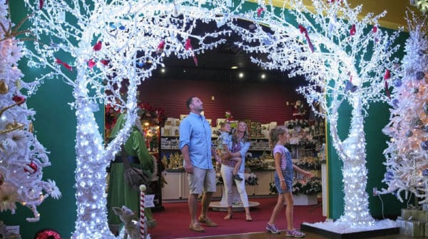 A father, daughter, and mother holding her son walk under the branches of artificial white lighted trees inside Santa Claus Christmas Store