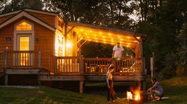 Cabin at Lake Rudolph is shown lit up at night with a person standing on the deck overlooking two people on either side of a campfire