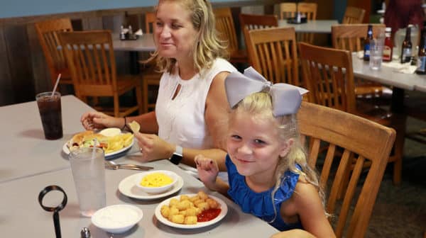 A mother and daughter sit at a restaurant table with meals in front of them, the daughter leaning toward the camera with a smile