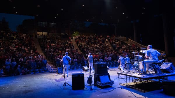 A band is shown from behind performing onstage at Lincoln Amphitheatre with a packed audience watching in the darkened background