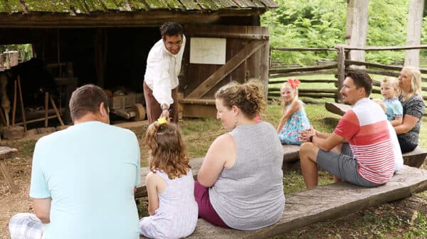 A ranger in period clothing interacts with a seated group in front of a log structure at the Living Historical Farm inside Lincoln Boyhood National Memorial