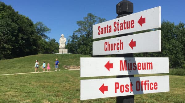 White directional signs with red lettering point to features at the Santa Claus Museum & Village while visitors walk past the historic Santa Claus statue in the background