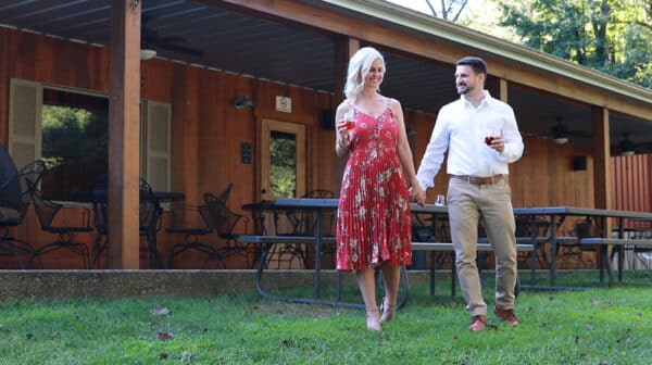 Woman in red floral dress and man in white dress shirt and khaki pants hold hands and carry wine glasses as they walk through the grass outside Pepper's Ridge Winery