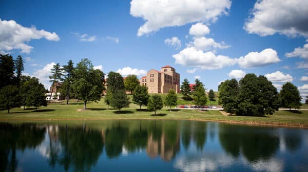 A large stone building at Saint Meinrad Archabbey is shown in the background with trees in front and a lake reflecting the trees in the foreground