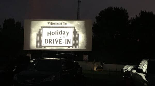 Cars are parked in front of a movie screen at Holiday Drive-In Movie Theater is lit up with the logo.