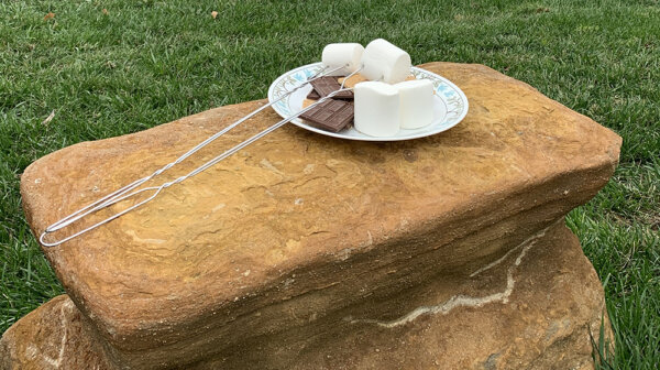 Plate with marshmallows, graham crackers, and chocolates and roasting sticks set on a sandstone rock