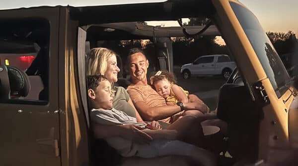 Mom and dad sit in front seat of Jeep with sleeping kids in their laps at Holiday Drive-In during destination video shoot