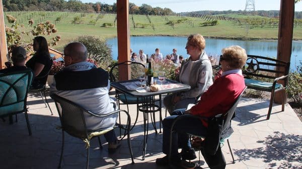 Visitors to Monkey Hollow Winery and Distillery sit at tables on the patio with lake and vineyard in the background