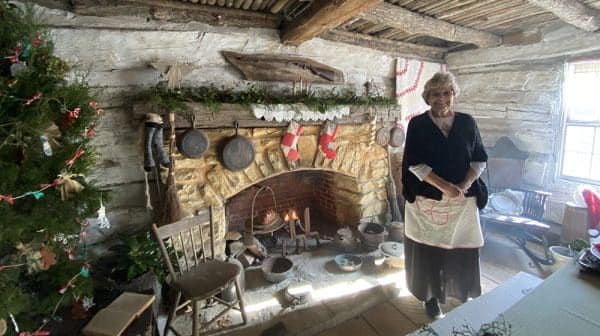 Woman in pioneer clothing stands in front of a fireplace inside a cabin at Lincoln Pioneer Village & Museum during the Treemendous Christmas in the Village